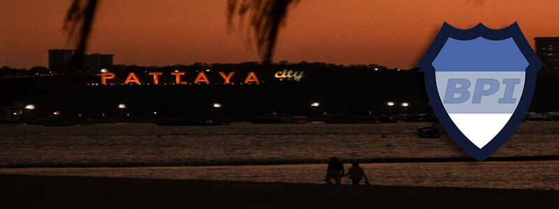 Two people walking on the beach at night with the Pattaya sign illuminated in the background and the BPI logo